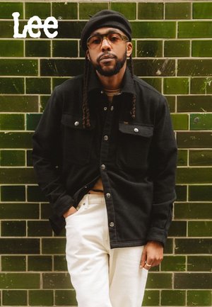 Schwarze Knopfleiste-Jacke aus Stoff mit strukturierter Oberfläche, getragen über einem hellen Top, kombiniert mit weißen Hosen vor einer grün gefliesten Wand.