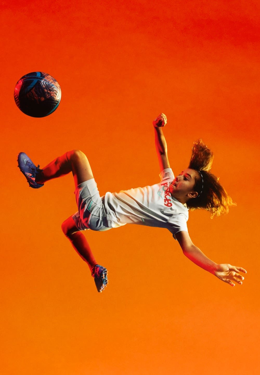 A person in a white T-shirt and gray shorts leaps, kicking a blue and black soccer ball against an orange background.