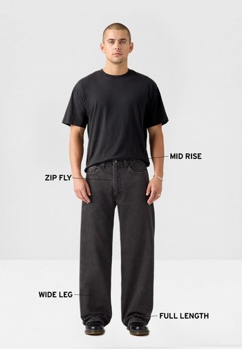 Hombre vistiendo jeans negros de pierna ancha con talle medio, cierre de cremallera y longitud completa, combinado con una camiseta negra lisa y zapatos negros sobre un fondo blanco.