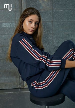 Jeune femme aux longs cheveux portant un survêtement bleu marine avec des rayures rouges et blanches, assise de côté sur un tabouret noir contre un mur gris.