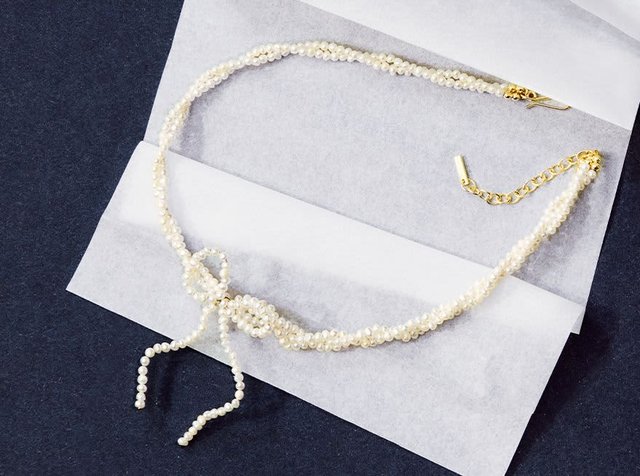 Λεπτή κολιέ με μαργαριτάρια και κόμπους, με χρυσό κούμπωμα, σε παρουσίαση πάνω από λευκό χαρτί περιτυλίγματος σε αντίθεση με σκούρο φόντο.