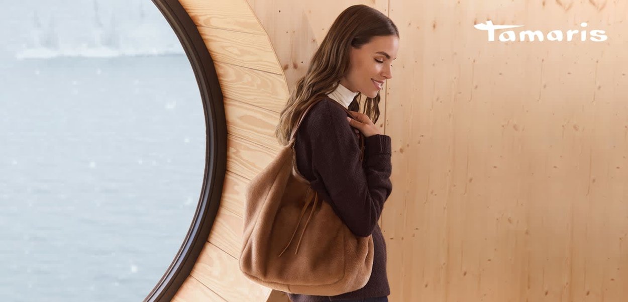 Braune, weich strukturierte Handtasche mit abgerundeter Form und Kordelzugverschluss, getragen über einem dunklen Pullover vor einem Hintergrund aus Holz und Wasser.