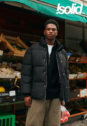 Giacca a vento nera imbottita con collo alto, chiusura con zip e tasche laterali. Il modello tiene una borsa trasparente con prodotti. Lo sfondo mostra un mercato.