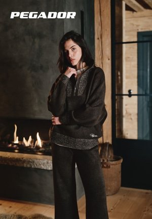 Frau in dunkler, gemütlicher Freizeitkleidung steht drinnen neben einem brennenden Kamin mit Holzwänden und Tür im Hintergrund.