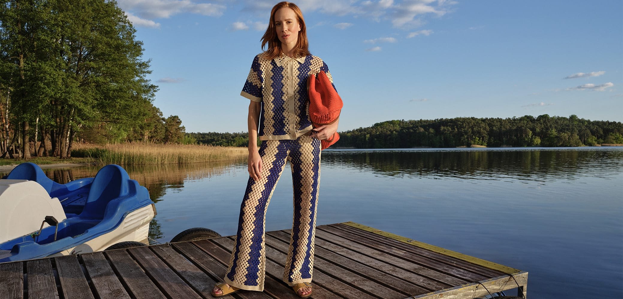 Háčkovaný dvoudílný outfit s geometrickým vzorem v modré a krémové barvě, doplněný živou oranžovou taškou, stojící na dřevěném mole u vody.