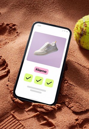 White athletic sneaker displayed on a phone screen, set against a sandy background. Phone shows payment options with checkmarks.