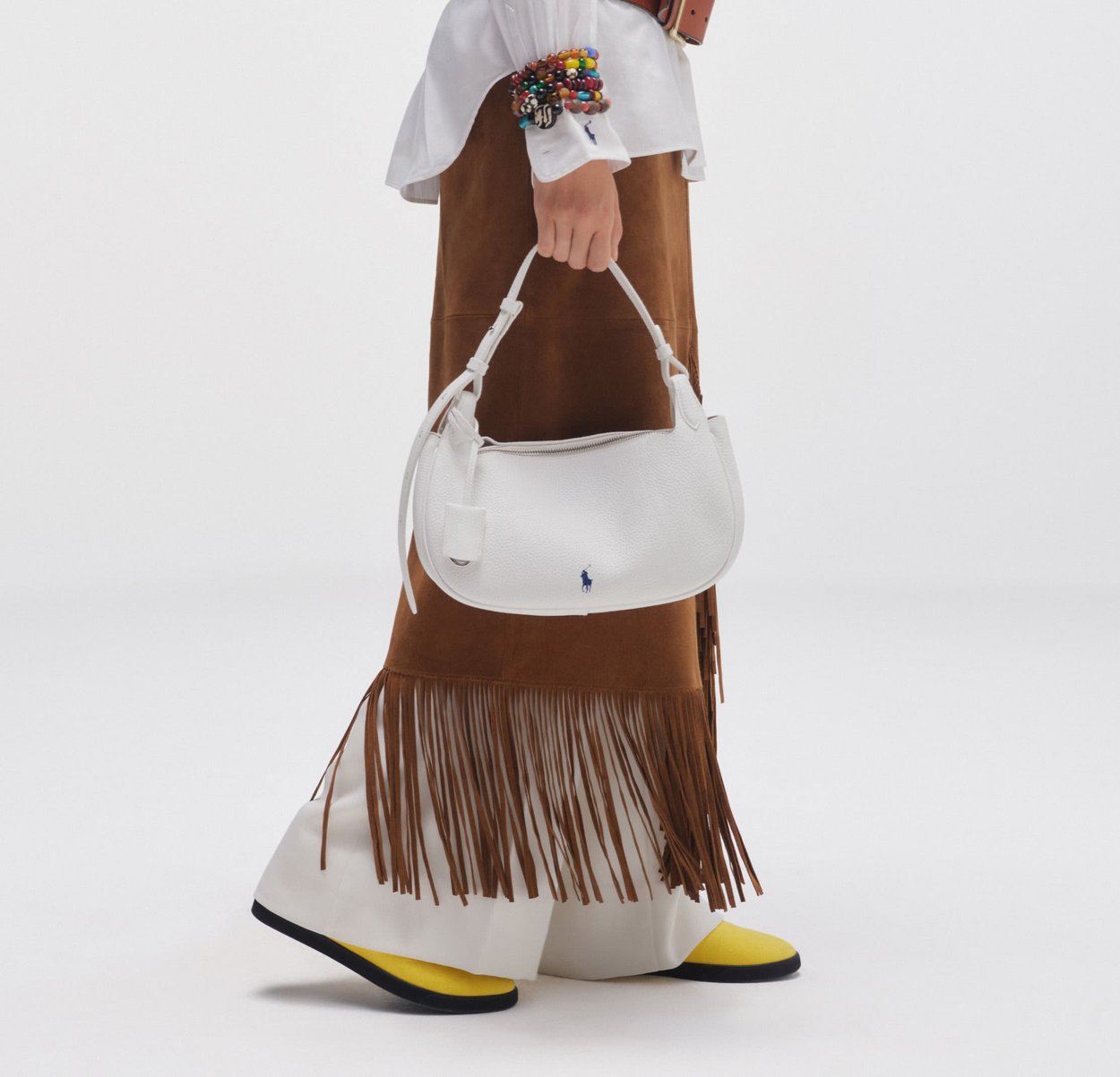 Persoon draagt een wit overhemd, bruin franjerokje, witte broek, gele schoenen, kleurrijke armbanden en houdt een witte leren handtas met een blauw logo vast.