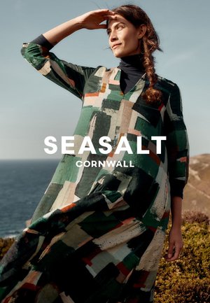 Vrouw in een gebloemde jurk die haar ogen beschut, staand op de klif met uitzicht op de oceaan en een heldere lucht op de achtergrond.