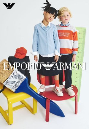 Due bambini posano con accessori e sedie colorate. Si vedono un pennello blu e un righello. Il ragazzo indossa una felpa a righe arancione e bianca.