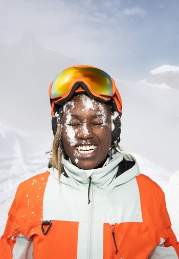 Skijacke in Hellblau mit leuchtend orangefarbenen Akzenten, trägt orangefarbene Skibrille; Schnee bedeckt Gesicht und Jacke in einer verschneiten Berglandschaft.