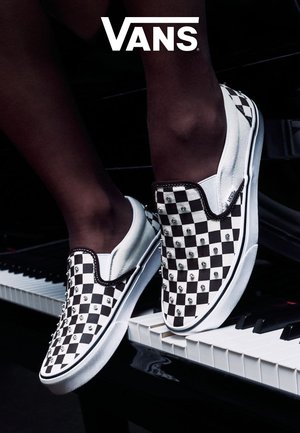 Pies con zapatillas Vans slip-on a cuadros en blanco y negro con tachuelas de metal descansando sobre un teclado de piano, logo de Vans arriba.