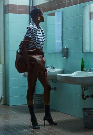 Mujer de cabello corto y grandes pendientes está de pie sosteniendo un bolso grande marrón en un baño con azulejos frente a dos lavabos con espejos.