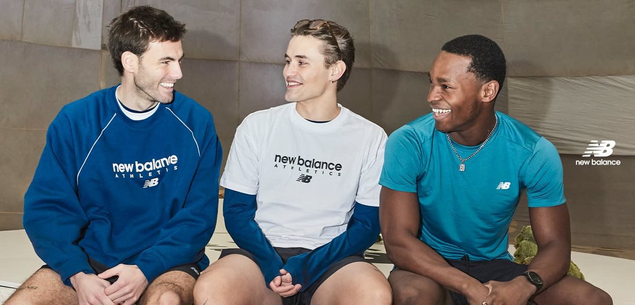 Drie jonge mannen in New Balance sportkleding zitten en lachen, terwijl ze een vriendelijk gesprek voeren tegen een neutrale achtergrond.