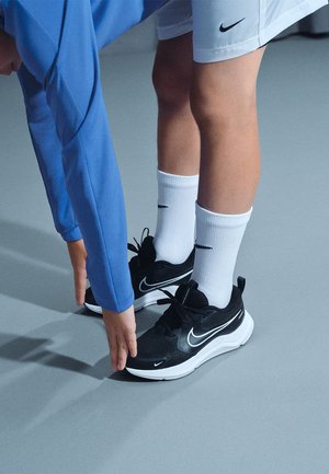 Baskets de sport noires avec des accents blancs, dotées d'un bout arrondi, d'une tige en mesh et d'une semelle rembourrée. Portées avec des chaussettes blanches et un short.