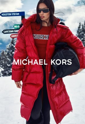 Abrigo largo rojo, acolchado, con acabado brillante, superpuesto sobre una sudadera roja con texto blanco. La modelo sostiene un bolso negro, ambientada en un paisaje nevado.