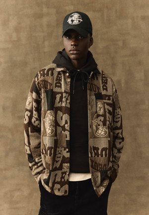 Jonge man met een bruin patroonjasje over een zwarte hoodie, zwarte pet met globe-logo, staand tegen een bruine, getextureerde achtergrond.