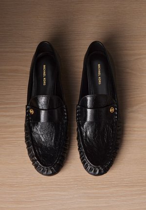 Par de mocasines de cuero negro de Michael Kors con emblema dorado MK sobre superficie de madera, vistos desde arriba.