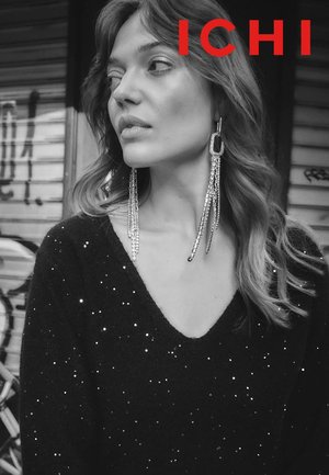 Schwarzer Pullover mit V-Ausschnitt, verziert mit kleinen silbernen Pailletten. Lange, funkelnde Ohrringe mit geometrischem Design. Im Hintergrund ist Graffiti zu sehen.