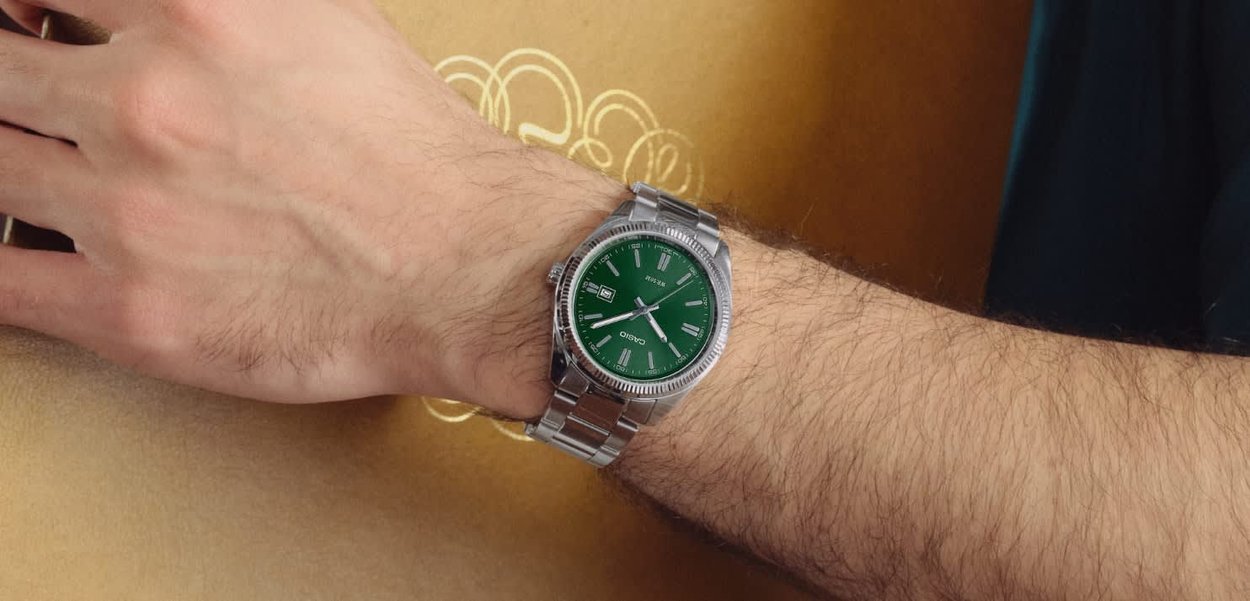 Pols met een zilveren metalen horloge met een groene wijzerplaat die de tijd 10:09 en de datum 20 toont op een goudkleurige patroonoppervlakte.