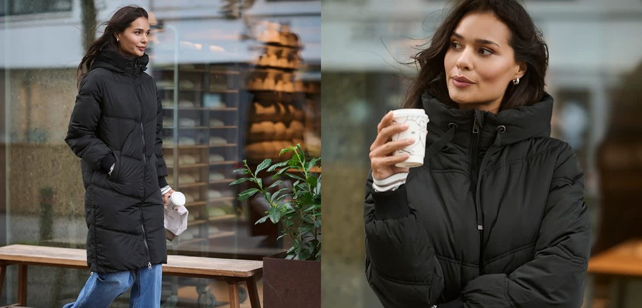 Svart dunjacka med huva, med quiltad design, dragkedja och sidofickor. Bärs med blå jeans. Håller en kaffekopp.