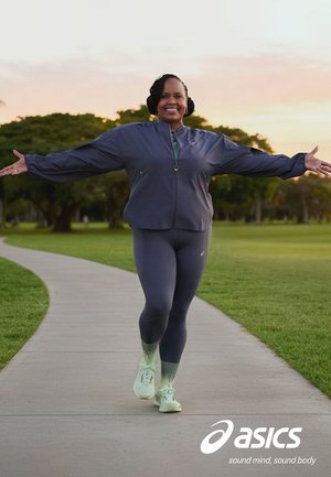 Lachende vrouw in sportkleding wandelt met uitgestrekte armen over een gebogen parkpad tijdens zonsondergang, met lichte groene hardloopschoenen aan.