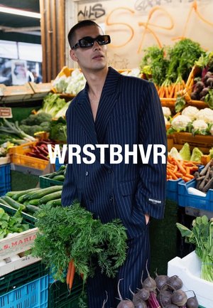 Osoba w granatowym garniturze w prążki, podwójnym zapięciu i czarnych okularach przeciwsłonecznych, trzymająca świeże marchewki wśród różnych warzyw na targowisku.