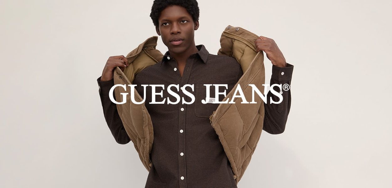 Ein junger Mann trägt ein kariertes Hemd und hebt eine braune gepolsterte Jacke vor einem einfachen beigen Hintergrund mit dem Textoverlay "GUESS JEANS".