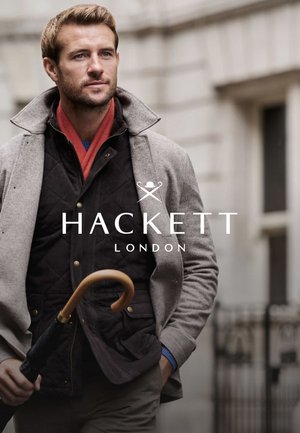 Man in een grijze wollen jas over een donker doorgestikte jas, met een houten paraplu in zijn hand. Draagt een rode sjaal en een blauwe trui eronder.