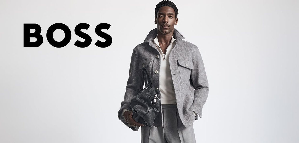 Graue Wolljacke mit Knopfleiste, zwei Brusttaschen, kombiniert mit einem weißen Hemd und grauen Hosen. Schwarze Lederhandtasche in der Hand.