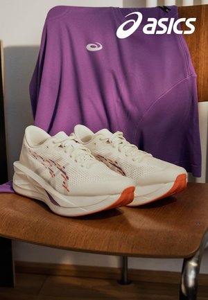 Des chaussures de sport blanches avec une tige texturée, des accents violets et orange, et une semelle rembourrée, à côté d'un t-shirt violet sur une chaise en bois.