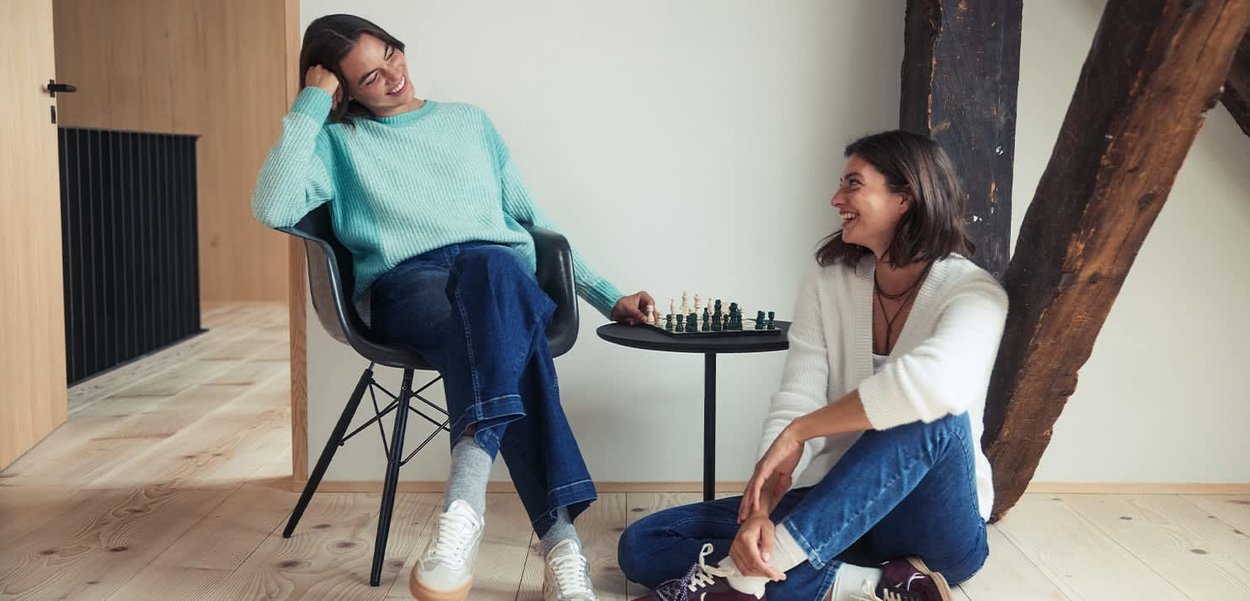 Zwei Personen in einem modernen Innenraum, eine sitzt auf einem schwarzen Stuhl, die andere auf dem Boden. Ein kleiner schwarzer Tisch hält ein Schachspiel bereit.