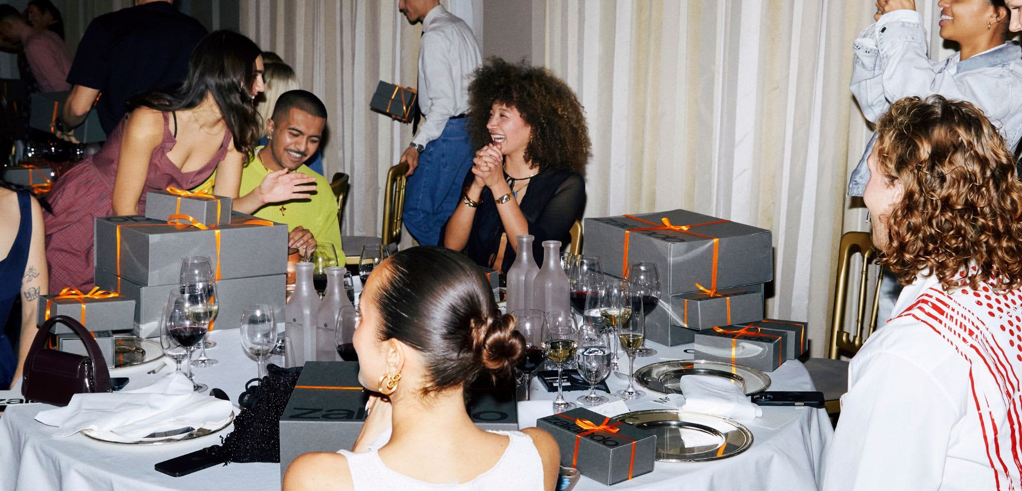 Et spisebord pyntet med grå bokse bundet med orange bånd, vinglas og hvide servietter, omgivet af forskellige gæster.
