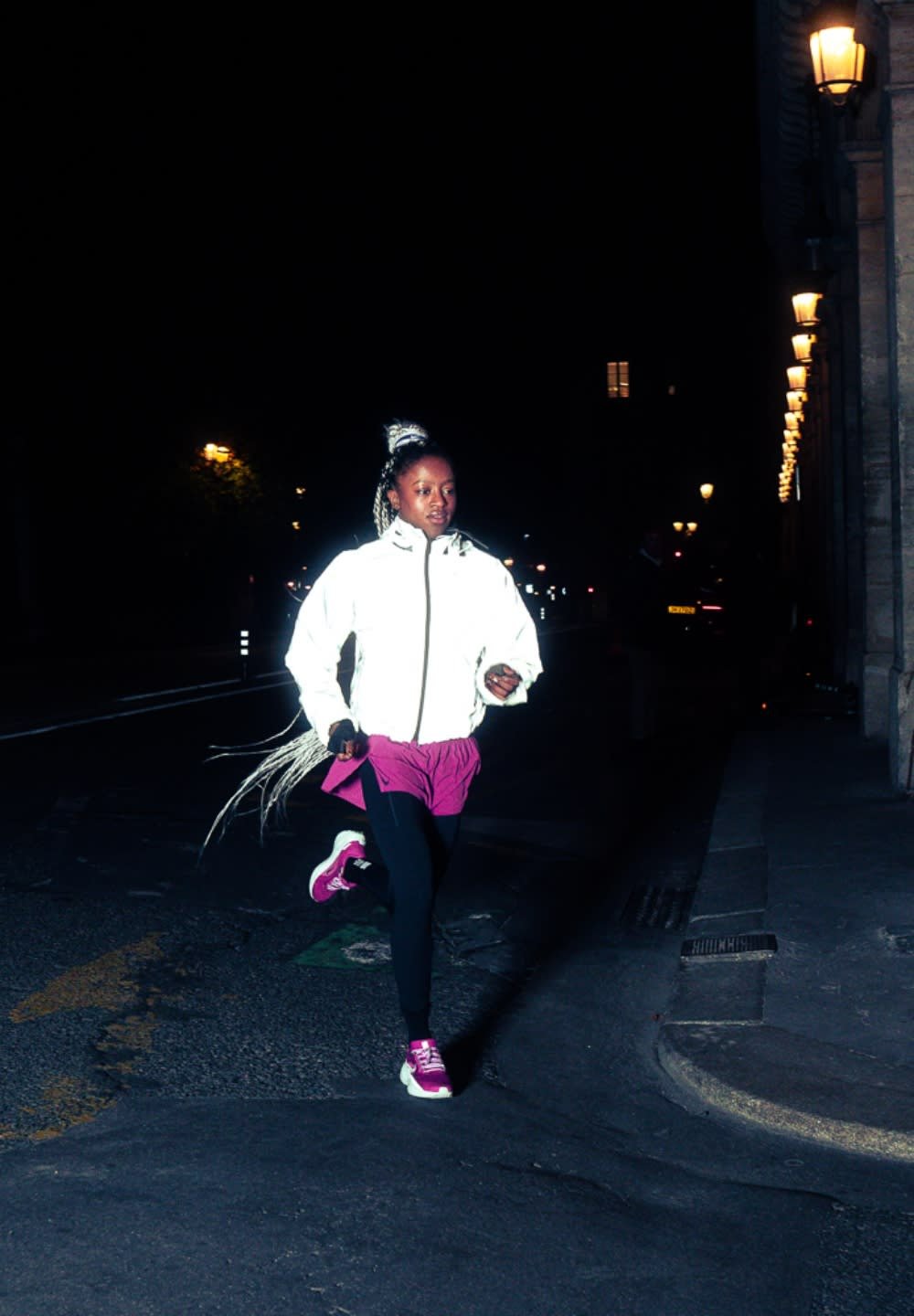 A person runs at night wearing a white reflective jacket, pink shorts, black leggings, and pink sneakers, with long braided hair.