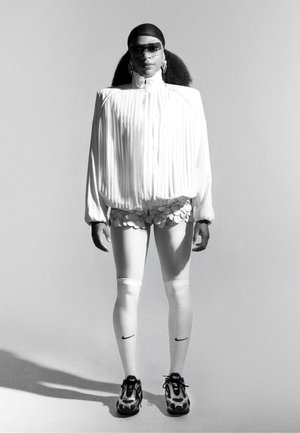 Witte geplisseerde jas, scalloped shorts, witte leggings en patronen sneakers. Accessoires bestaan uit zonnebril en creolen. Monochrome achtergrond.