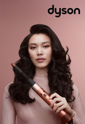 Femme aux longs cheveux ondulés et foncés tenant un boucleur à cheveux Dyson en métal rose doré sur un fond rose avec le logo Dyson.