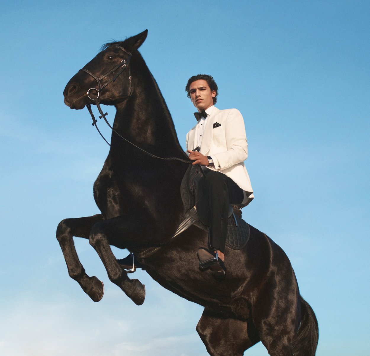 Hombre con esmoquin blanco monta un caballo negro encabritado contra un fondo de cielo azul claro.