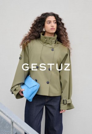 Vrouw met lang, krullend haar draagt een oversized olijfgroene jas en houdt een felblauwe bontclutch vast tegen een effen lichtgrijze achtergrond.