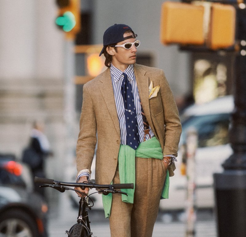 Giovane uomo che indossa un completo beige, camicia a righe, cravatta blu navy, occhiali da sole bianchi e cappellino, che cammina con la bici per strada in città.