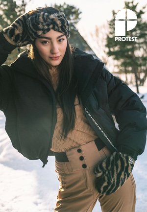 Jeune femme en plein air dans la neige portant une veste noire, un pantalon beige, des moufles à motifs et un bandeau assorti, avec des arbres en arrière-plan.