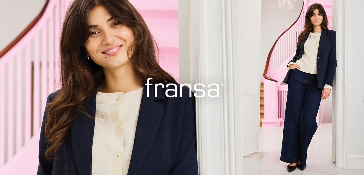 Junge Frau mit langen braunen Haaren, die einen marineblauen Blazer und weit geschnittene Hosen mit einer beigen Bluse trägt, posiert drinnen neben einem weißen Treppengeländer.