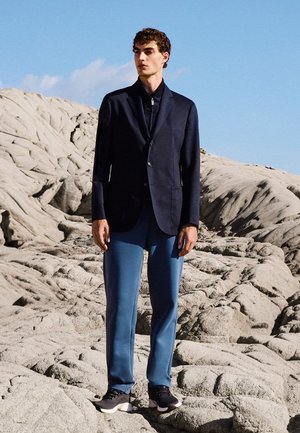 Jonge man met een marineblauwe blazer, zwart vest met rits, blauwe broek en zwarte sneakers staat op een rotsachtig terrein met textuur onder een heldere blauwe lucht.