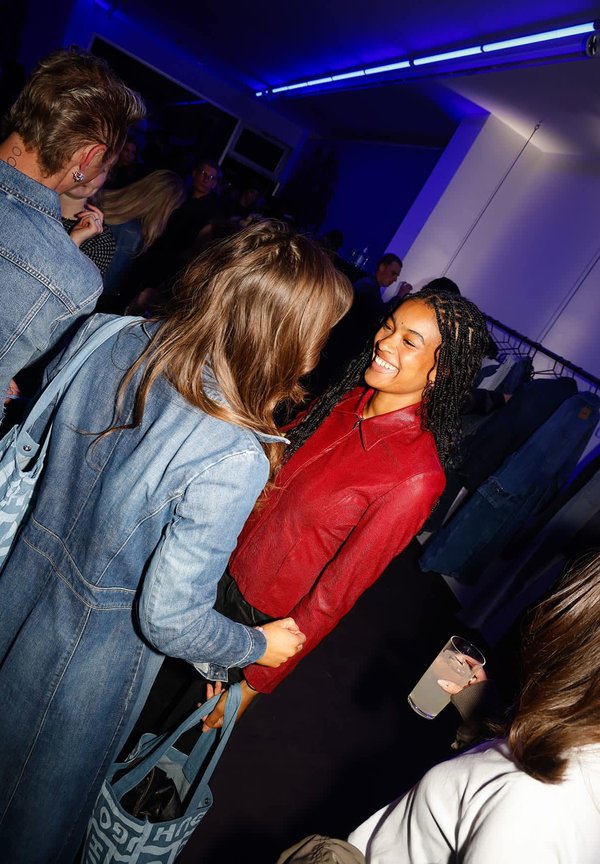 Two women are engaged in conversation. One wears a red leather jacket, the other a light blue denim jacket, against a dark backdrop.