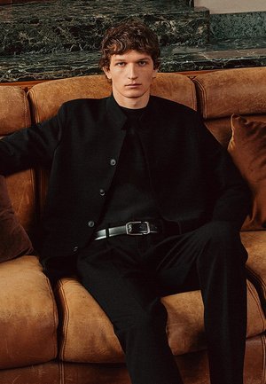 Veste noire à boutons sur un haut noir, associée à un pantalon noir et une ceinture. Le modèle est assis sur un canapé en cuir marron.