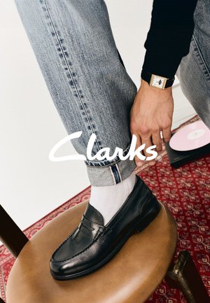 Black leather loafers with a smooth texture, worn with white socks, paired with cuffed blue jeans. Set against a patterned backdrop.