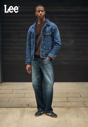 Denim jacket with a faded blue finish, brown knitted sweater, and blue jeans. Black shoes with white stripes. Gray background.