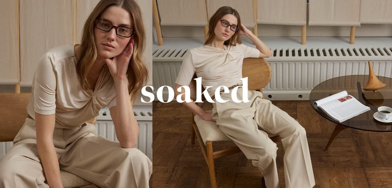 Frau mit Brille und beiger Kleidung sitzt auf einem Holzstuhl neben einem runden Glastisch mit einem Buch und einer Tasse in einem minimalistischen Raum.