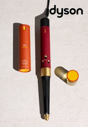 Ferro arricciacapelli Dyson con manico rosso e oro, spray condizionante leave-in arancione e piccolo contenitore con coperchio giallo su una superficie chiara.