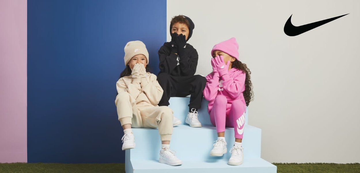 Troje dzieci siedzi na jasnoniebieskim stopniu. Niosą bluzy z kapturem oraz spodnie w kolorach beżowym, czarnym i różowym. Mają na sobie białe trampki i rękawiczki.