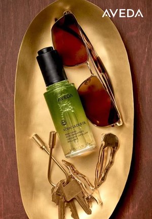 Aveda Miraculous Oil-flaske (grønt glas, sort låg) på en oval guldbakke, ledsaget af brune solbriller, messingnøglebundt og metalhårklips.
