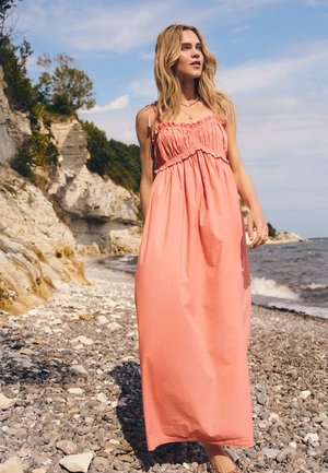 Vrouw met lang blond haar, gekleed in een koraalrode mouwloze maxi-jurk, staand op een rotsachtig strand met kliffen en zee op de achtergrond.